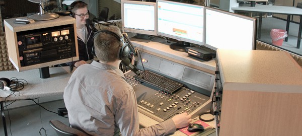 Ein junger Mann sitzt in einem Radiostudio an einem Mischpult und blickt auf Computermonitore
