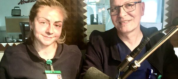 Zwei Beschäftige in der Krankenhaus-Hygiene sitzen in Dienstkleidung vor einem Mikrofon